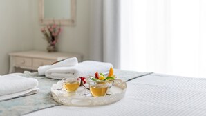 Un letto rifatto con asciugamani bianchi, un vassoio con due tazze di tè e frutta, e un vaso con fiori.