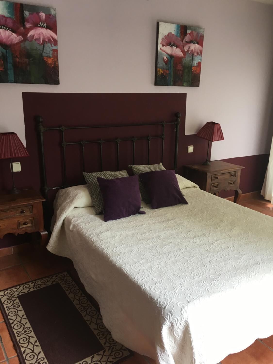 Un dormitorio con una cama, dos mesitas de noche, una lámpara roja, una mesita de noche de madera y una alfombra estampada.