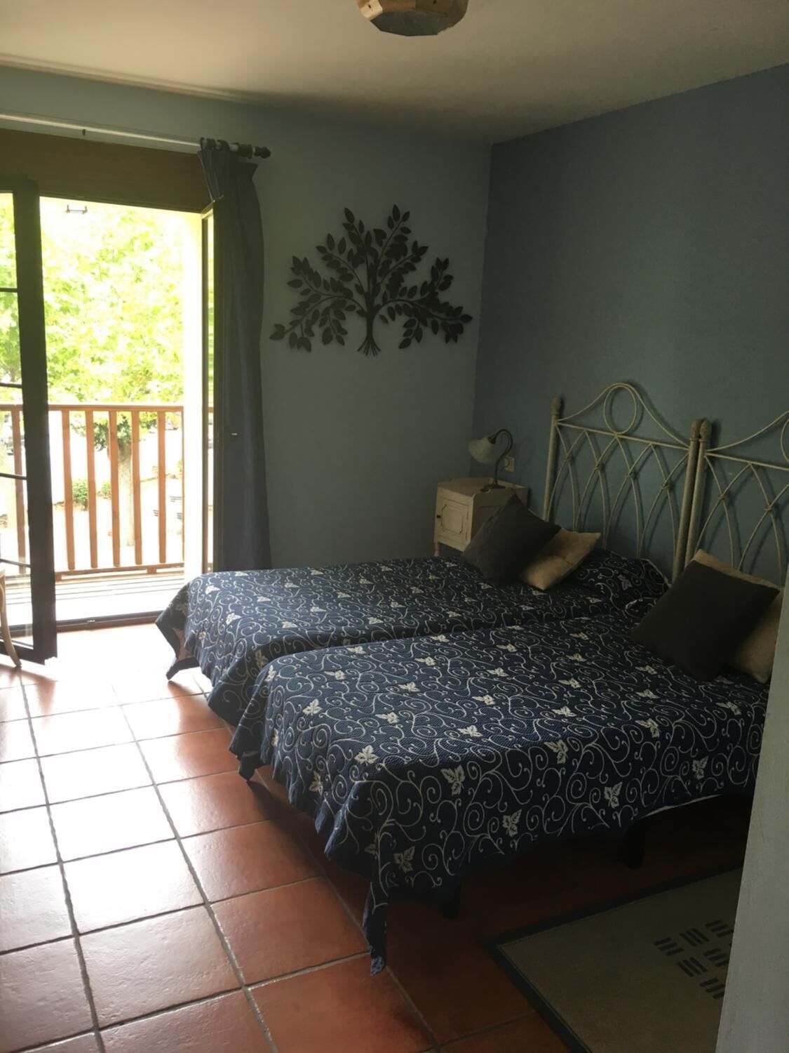 A bedroom with two beds, a small bedside table with a lamp, a wall-mounted tree decoration, and a view of the outdoors through a sliding door.