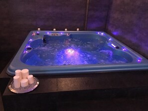 Una bañera de hidromasaje rectangular con iluminación azul, rodeada de velas, y una pequeña mesa redonda con velas.