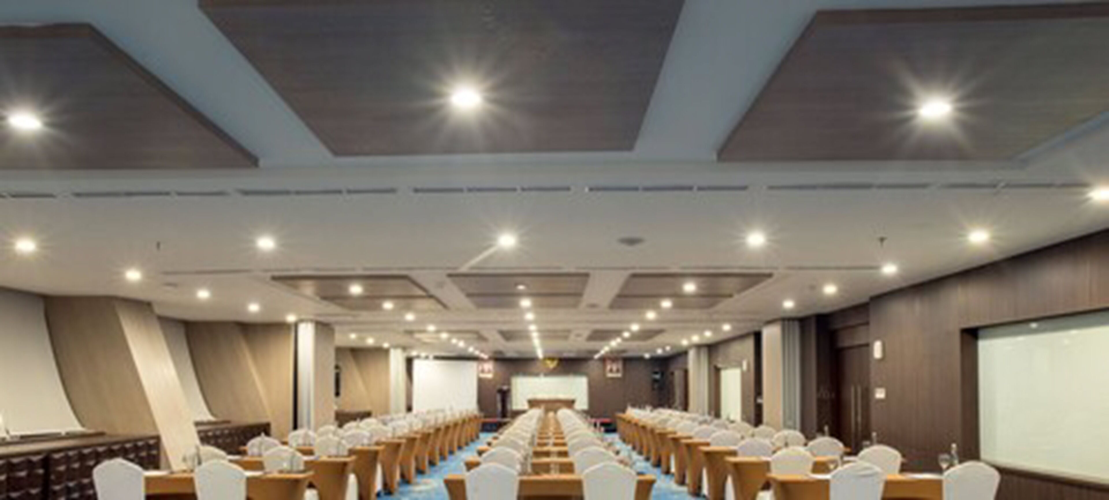 Une salle de conférence avec des rangées de tables et de chaises, un pupitre et un grand écran à l’avant.