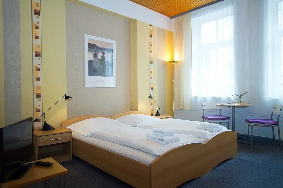 A hotel room with a double bed, bedside tables, a desk, a chair, and a window with curtains.