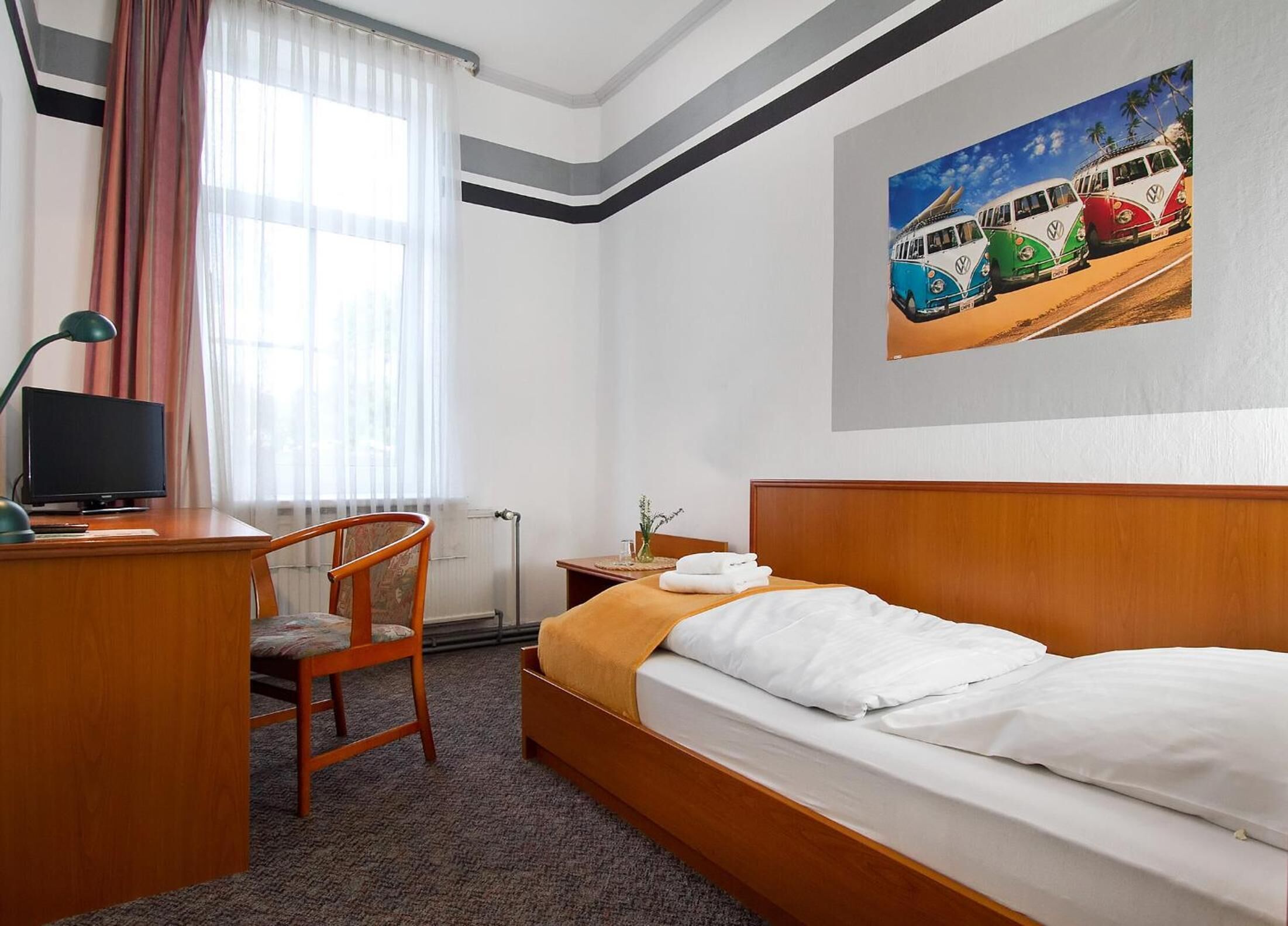 A hotel room with a wooden bed, a desk with a chair, a TV, and a painting of vintage buses on the wall.