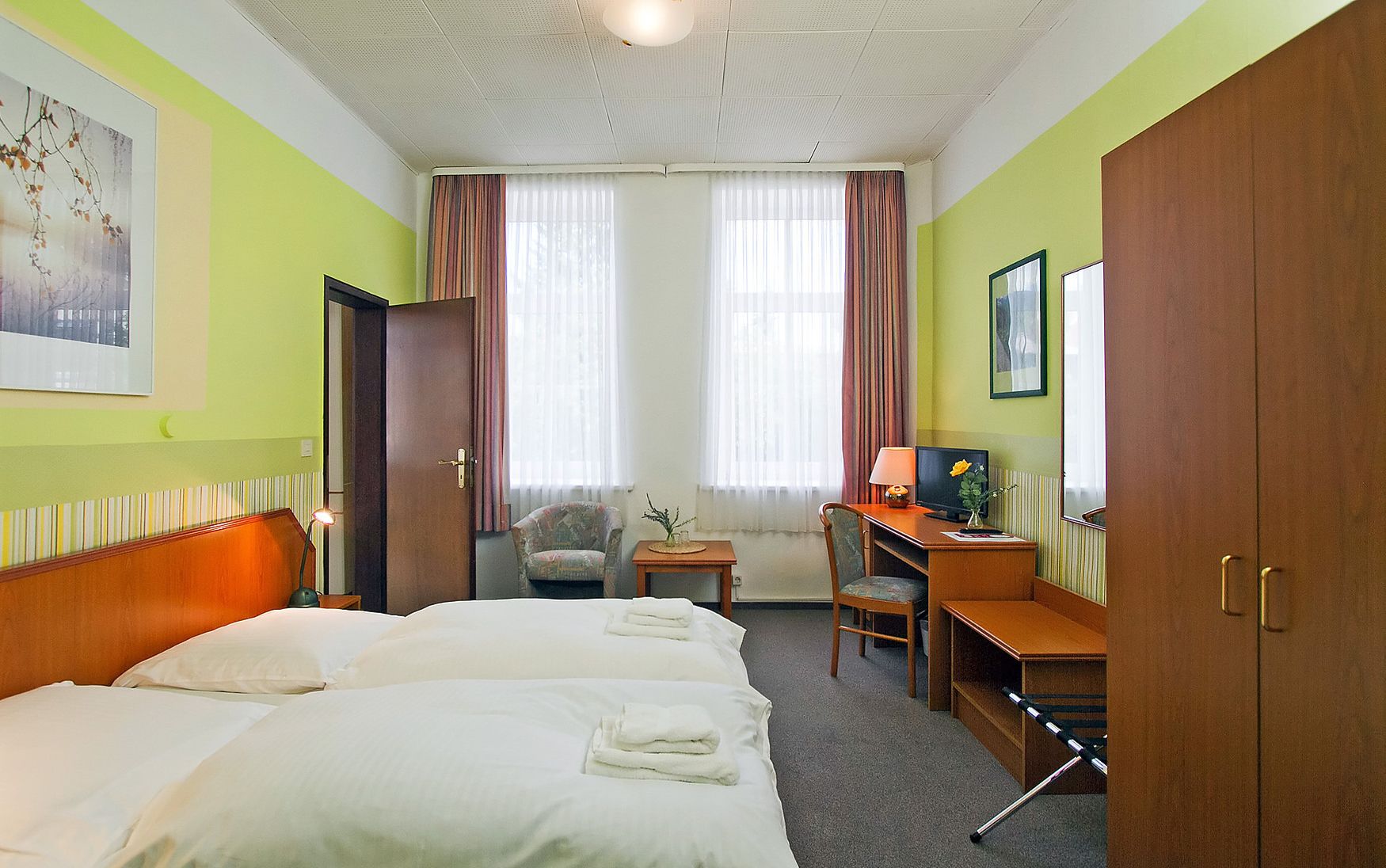Habitación de hotel con dos camas, un escritorio con una computadora, una silla y un ventanal con cortinas.