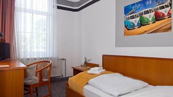Habitación de hotel con cama, escritorio, silla, radiador, ventana con cortinas y un cuadro colgado en la pared que representa una furgoneta.