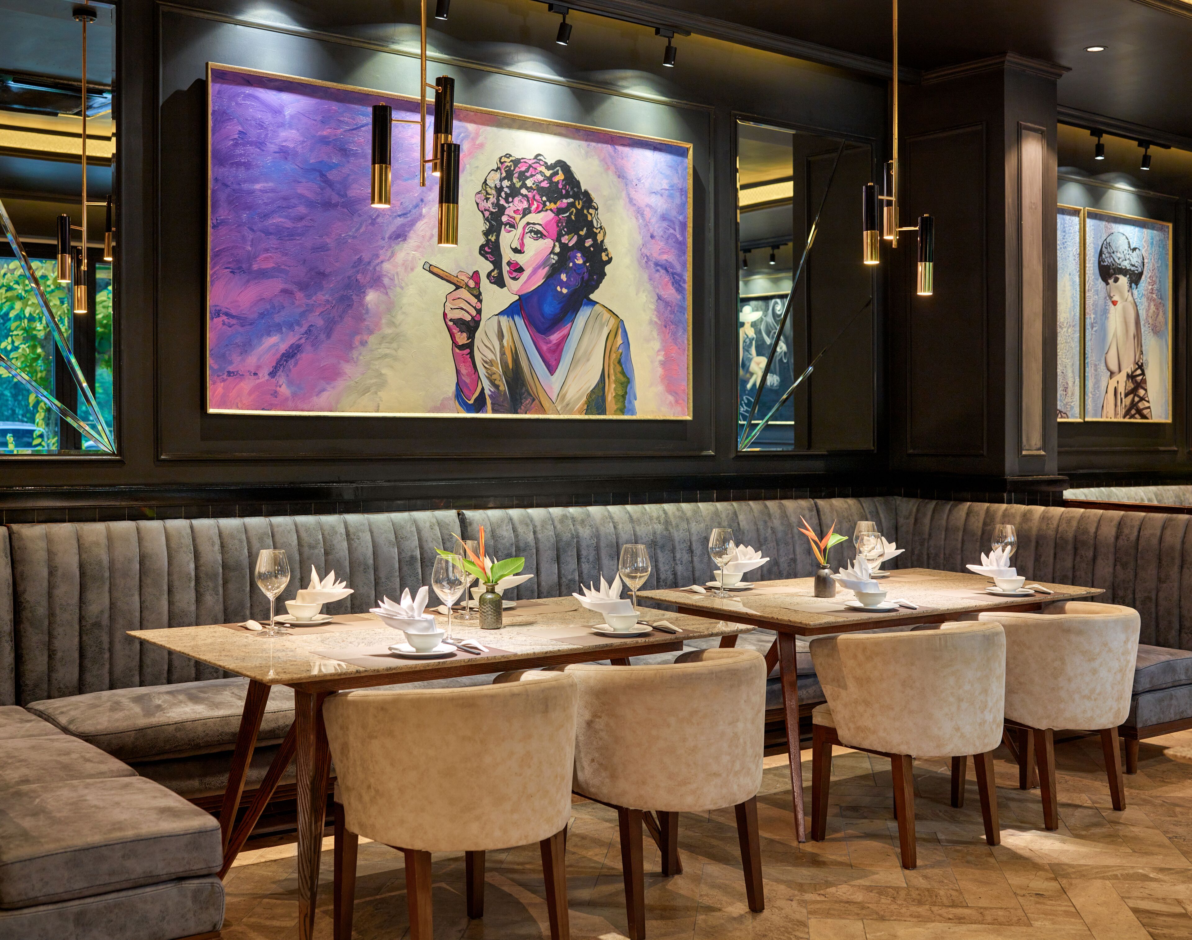 A restaurant with a modern interior, featuring a large mural, cushioned seating, and a wooden table with a vase of flowers.