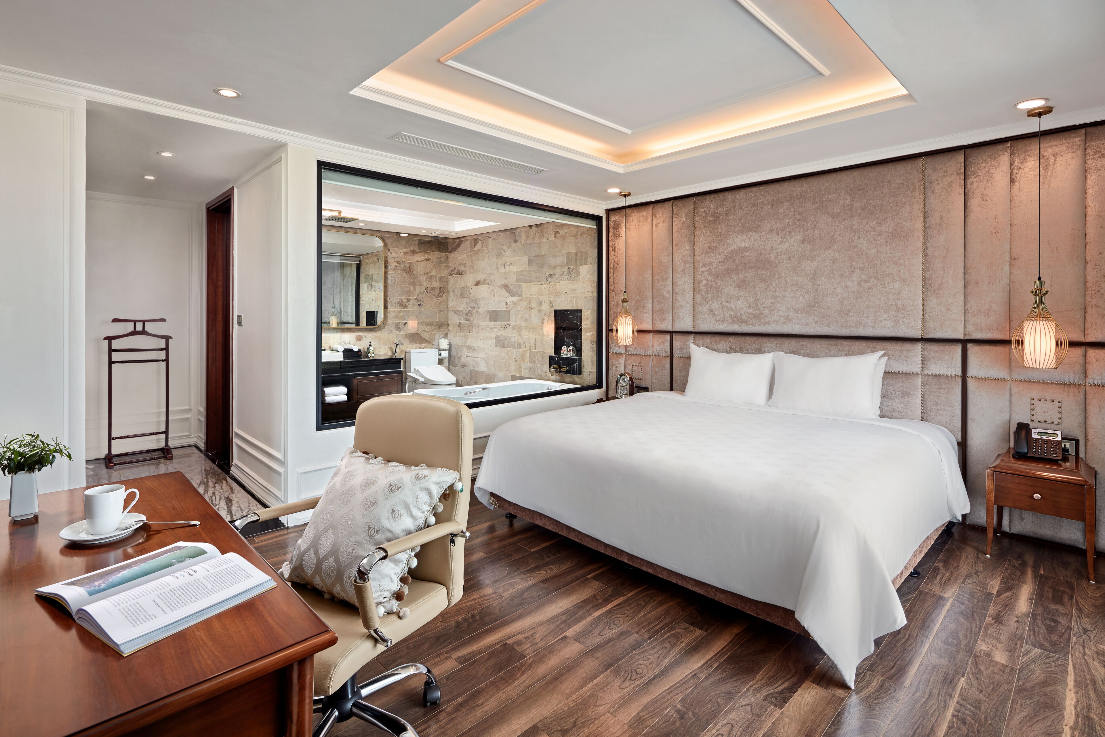 A modern hotel room with a large bed, a desk with a chair, a small table with a cup and saucer, and a large mirror.