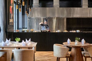 A restaurant with a chef preparing food at a counter, tables set with white napkins and plates, and a modern interior design.