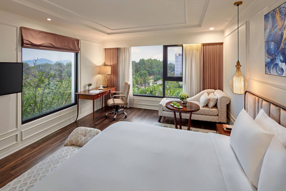 Quarto de hotel com uma cama grande, uma escrivaninha com cadeira, uma mesinha de apoio com abajur, e vista para o verde lá fora.