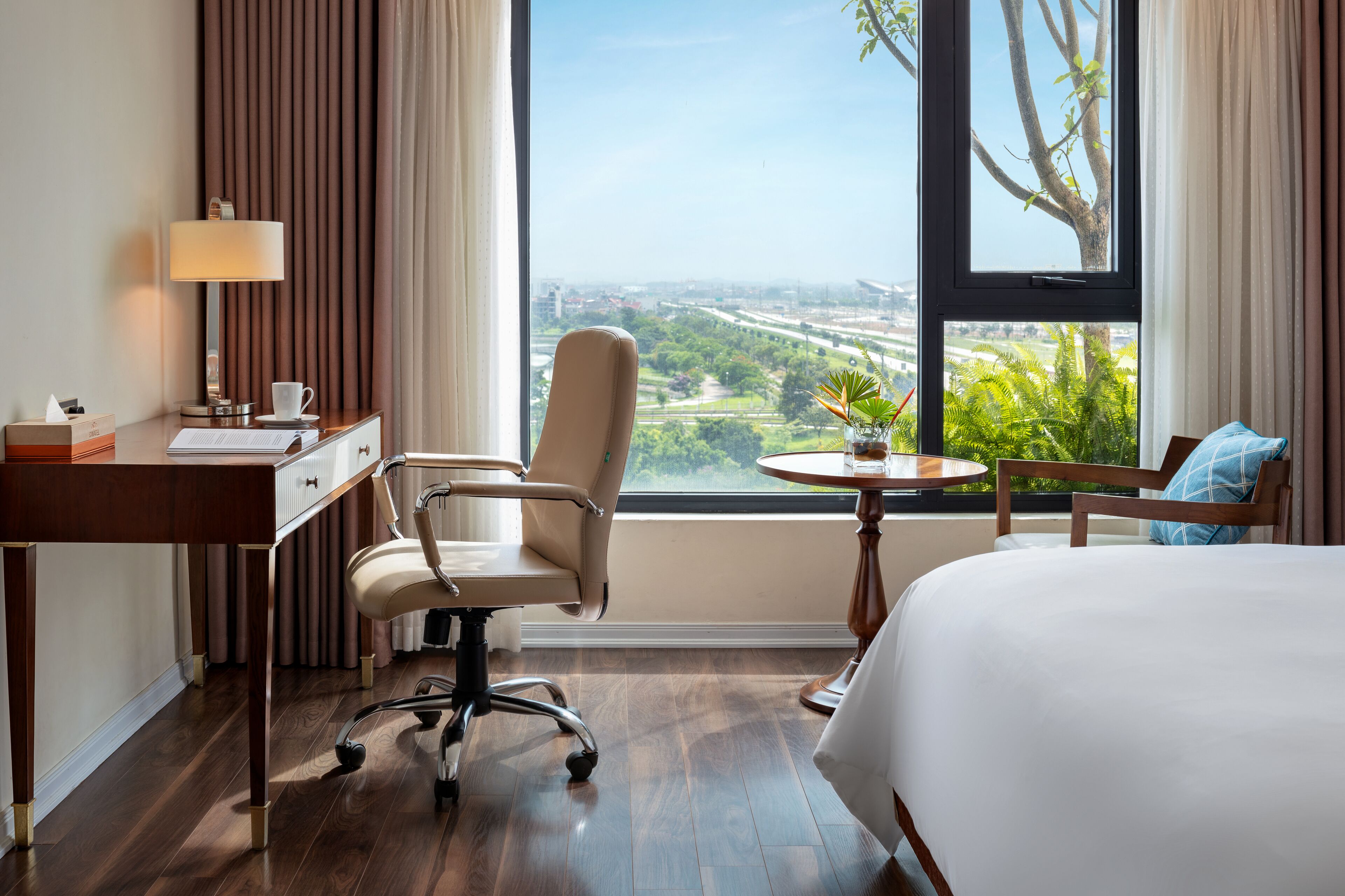 Une chambre d’hôtel équipée d’un bureau, d’une chaise, d’un lit et offrant une vue sur le paysage urbain grâce à de grandes fenêtres.