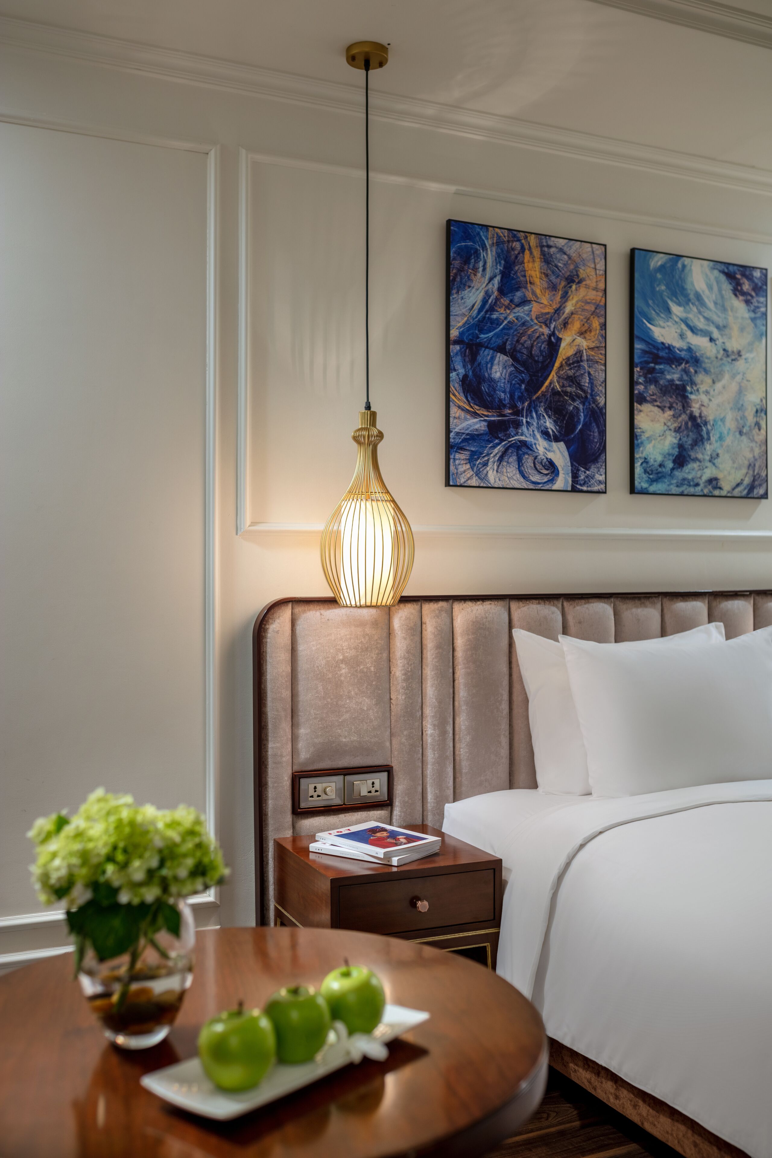 A hotel room with a bed, a bedside table with a vase of flowers and apples, a nightstand with a lamp, and abstract paintings on the wall.