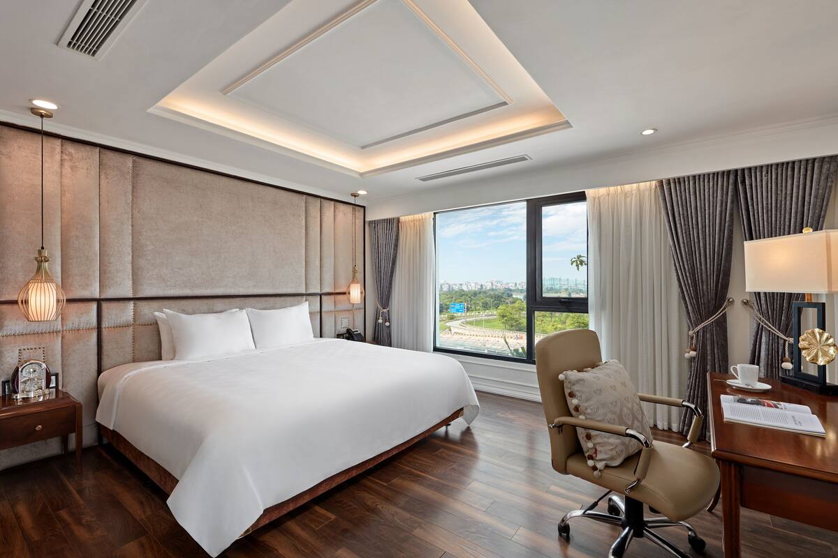 Quarto de hotel com uma cama grande, uma mesa e uma cadeira. Há uma janela com vista para a paisagem urbana.