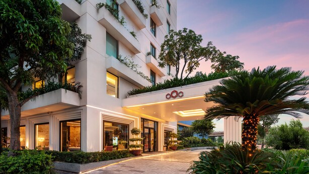 Una entrada de hotel moderna con fachada iluminada, zona de estar al aire libre y abundante vegetación.