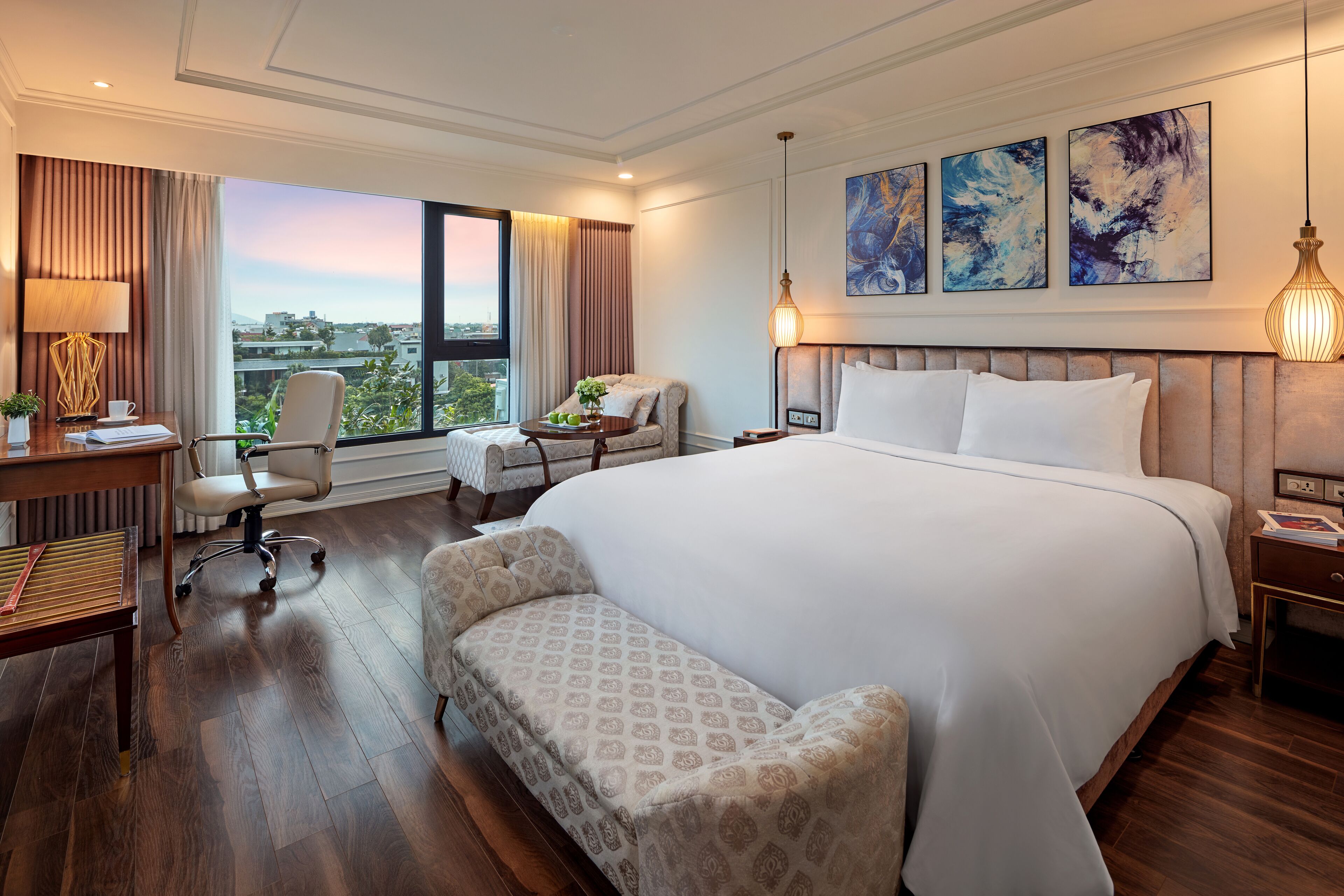 A hotel room with a large bed, a desk with a lamp, a chair, and a view of the city.
