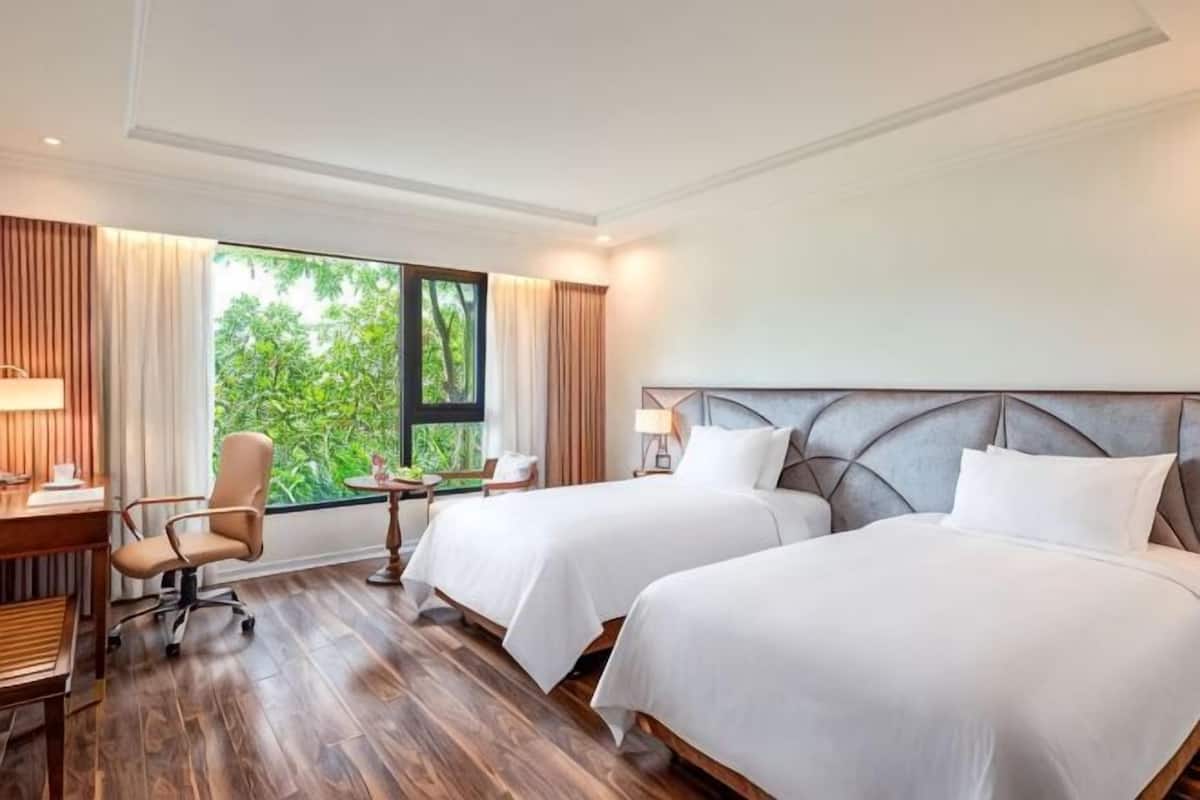 Quarto de hotel com duas camas, uma escrivaninha, uma cadeira e uma janela com vista para o verde.