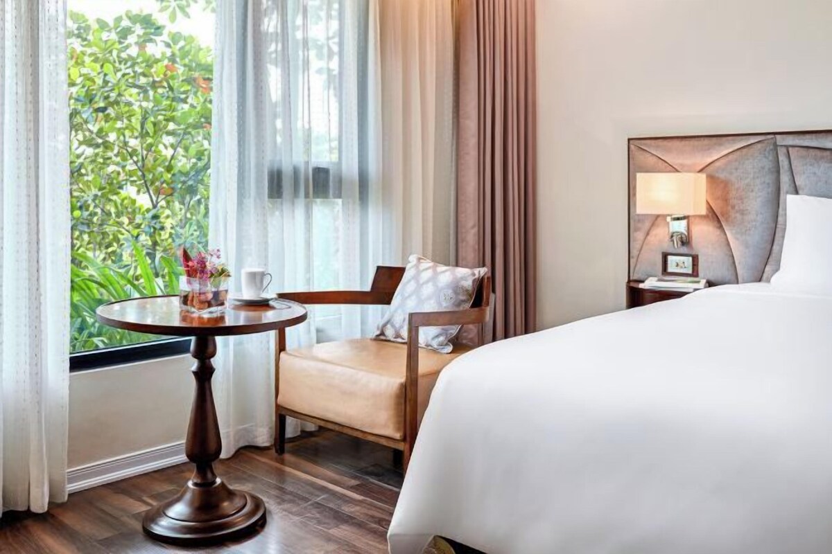 Una habitación de hotel con una cama, una silla, una mesita con una taza y un florero, y una ventana con cortinas.
