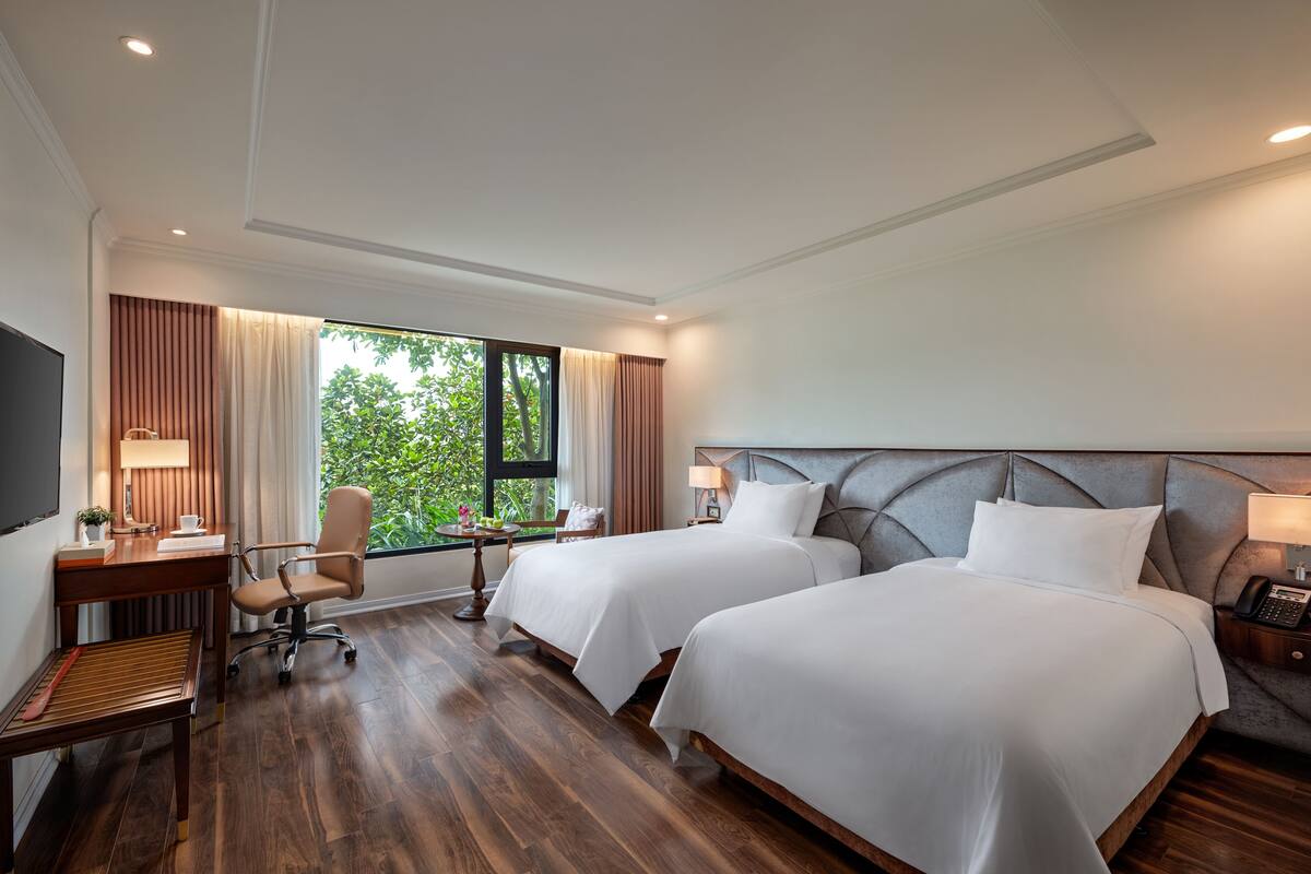 Quarto de hotel com duas camas, uma escrivaninha com cadeira, televisão e janela com cortinas.