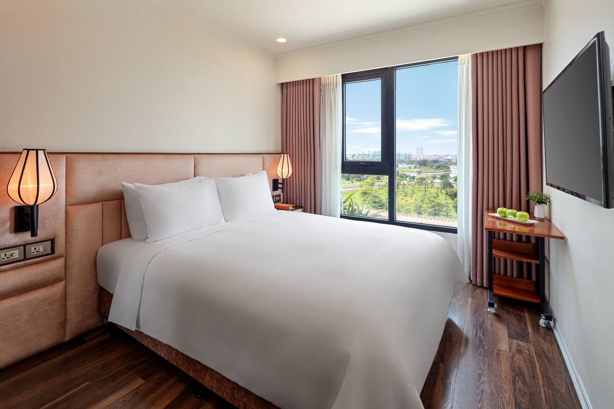 Quarto de hotel com uma cama grande, televisão, mesa de cabeceira, abajur, janela com vista e uma pequena mesa com frutas.