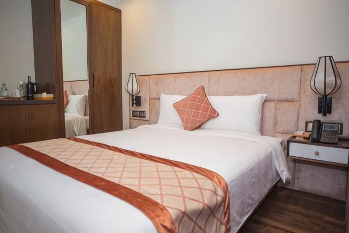 Habitación de hotel con una cama grande, una mesita de noche con lámpara, un espejo y un armario de madera.