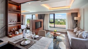 Una habitación de hotel moderna con zona de comedor, sofá, televisor y vista a la ciudad.