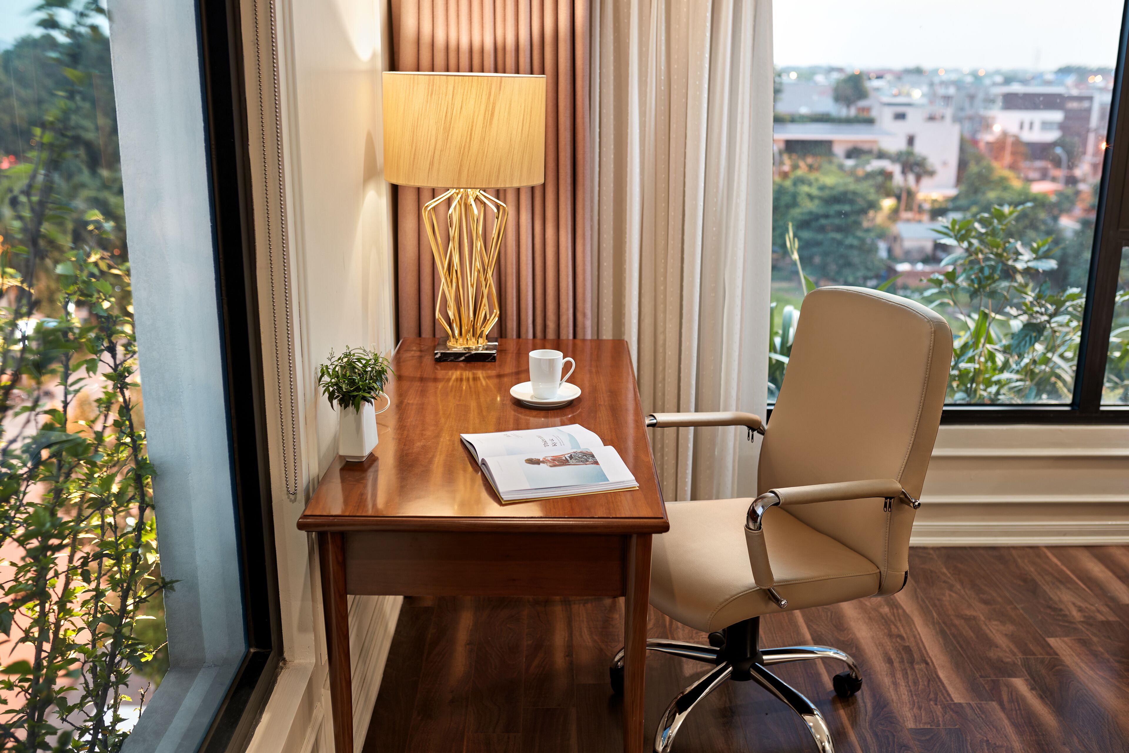 Un escritorio de madera con una lámpara, una taza y una revista, ubicado cerca de una ventana con vista a la ciudad.