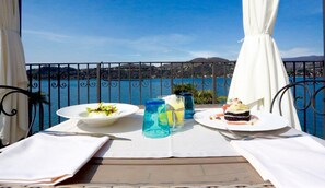 Een balkon met een tafel, uitzicht op zee, twee borden eten en twee glaasjes water.