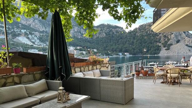A terrace with wicker furniture, a stone wall, and a view of a lake and mountains. - Es Apartments (Marmaris)