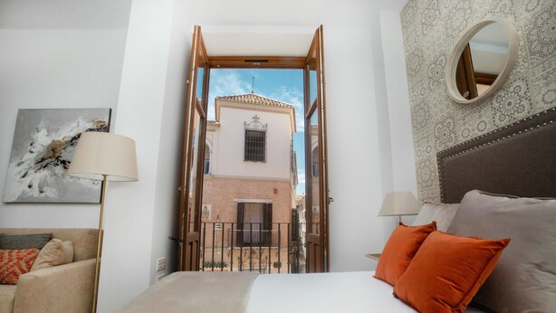 A bedroom with a bed, two pillows, a nightstand, a lamp, and a view of a building through an open door. - Feelathome Mártires Apartments (Málaga)