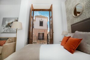 Une chambre à coucher avec un lit, deux oreillers, une table de chevet, une lampe et une vue sur un immeuble par une porte ouverte.