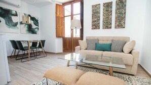 A modern living room with a glass coffee table, a beige sofa with cushions, a patterned rug, and a dining area with chairs and a glass table. - Feelathome Mártires Apartments (Málaga)