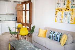 A modern kitchen with white cabinets, a dining area with a round table and chairs, and a living room with a sofa and cushions.