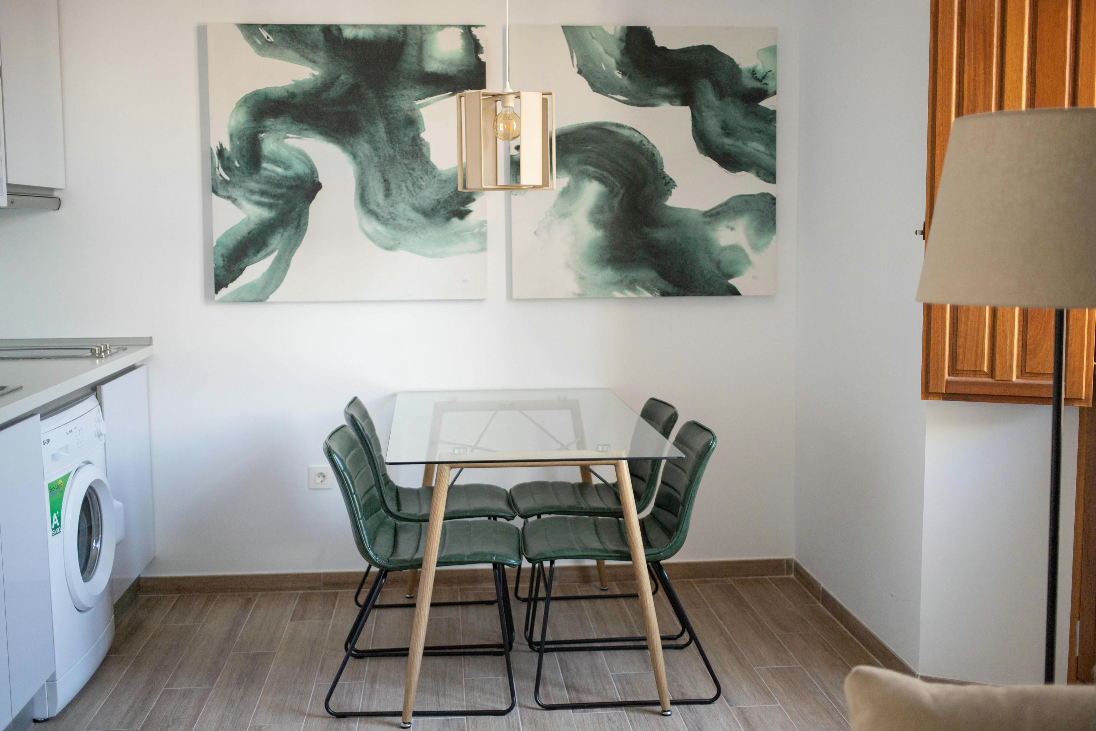 A modern kitchen with a glass dining table, green chairs, and abstract art on the wall.