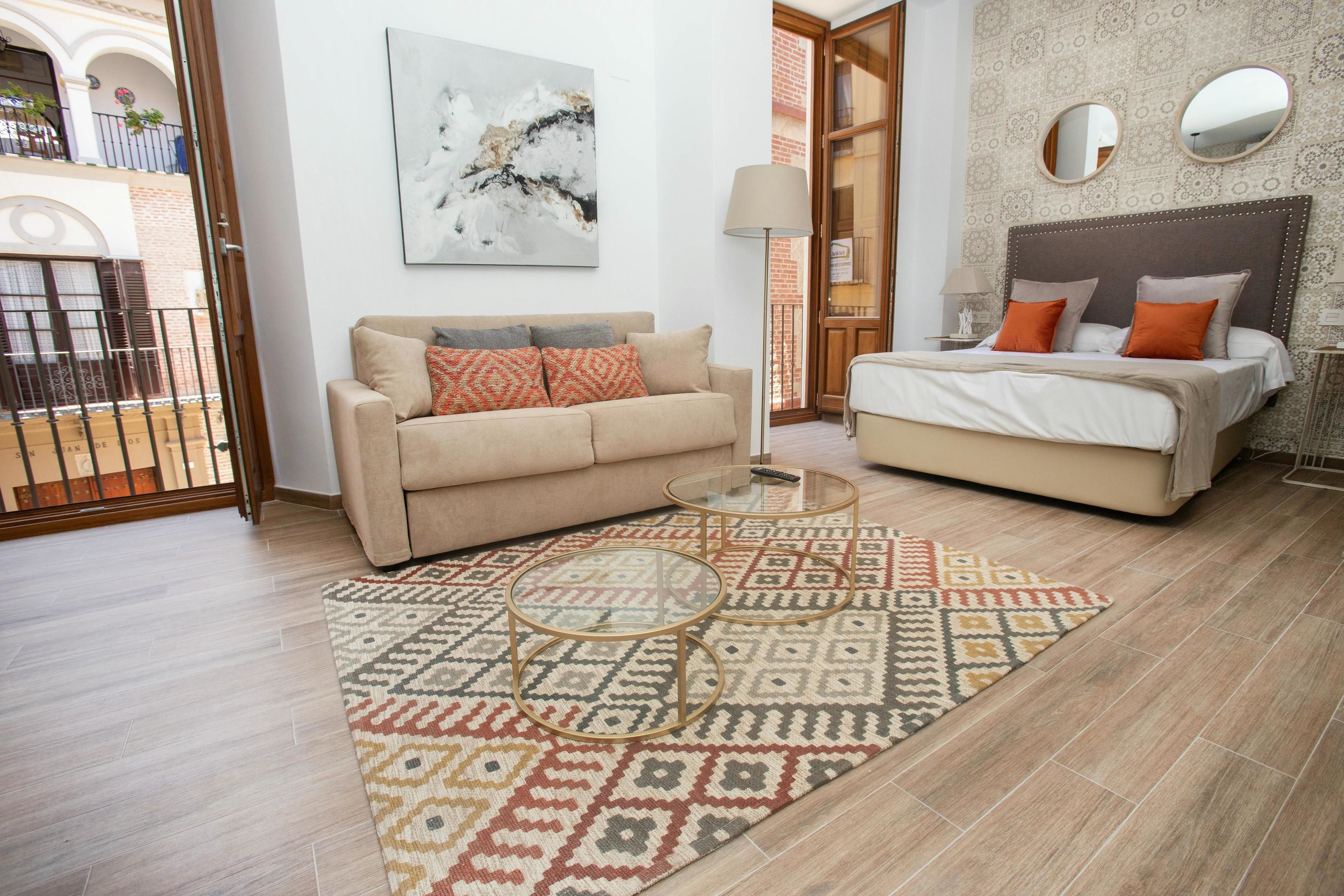 A modern bedroom with a beige sofa, a bed with orange pillows, a patterned rug, and a large mirror on the wall.