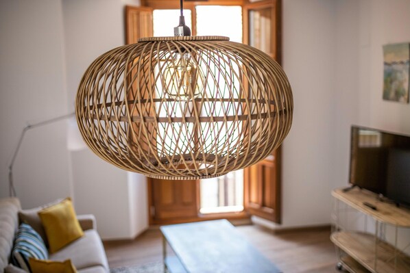 A room with a wicker pendant light, a sofa with cushions, a television on a wooden stand, and a wooden floor.
