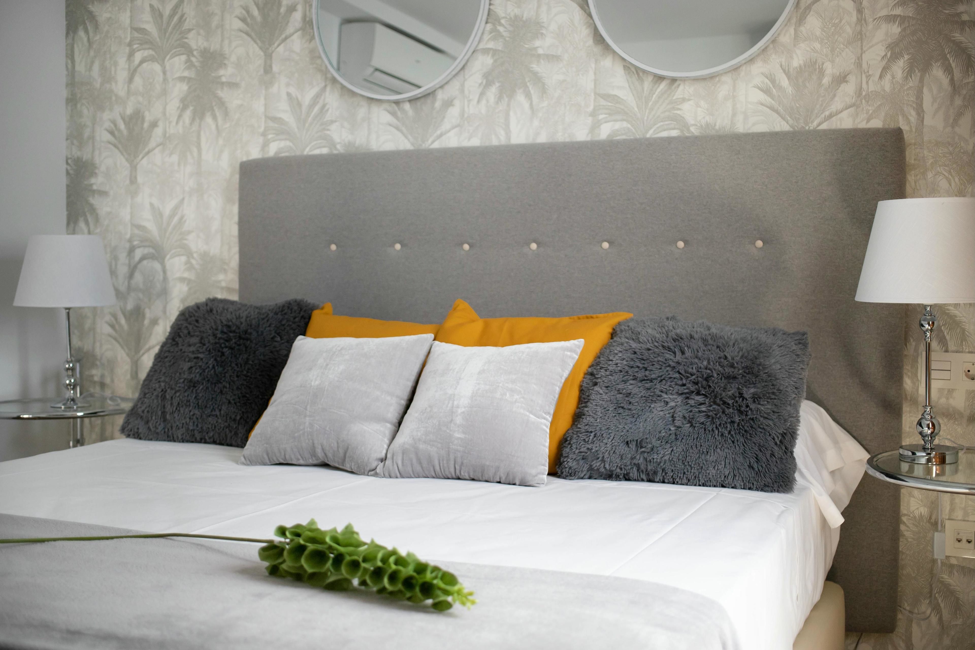 Una cama bien tendida con un cabecero gris, almohadas blancas y grises, y una planta verde.