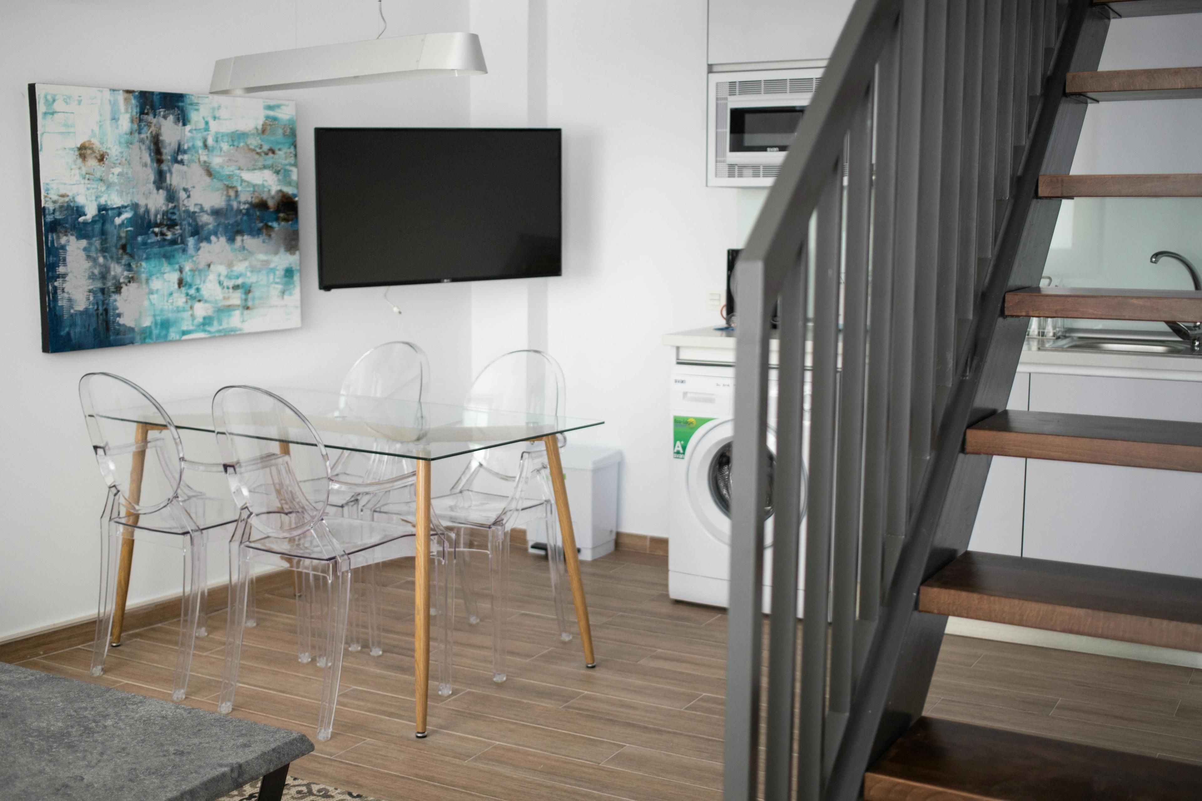 A modern kitchen with a washing machine, a dining area with a glass table and chairs, and a staircase.