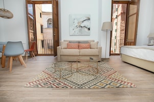 A living room with a beige sofa, a patterned rug, a small coffee table, a chair, and a bed.