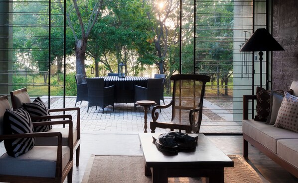 Un salón amplio con vistas a un jardín a través de ventanas grandes. Hay un sofá, una silla y una mesita con una cesta encima.