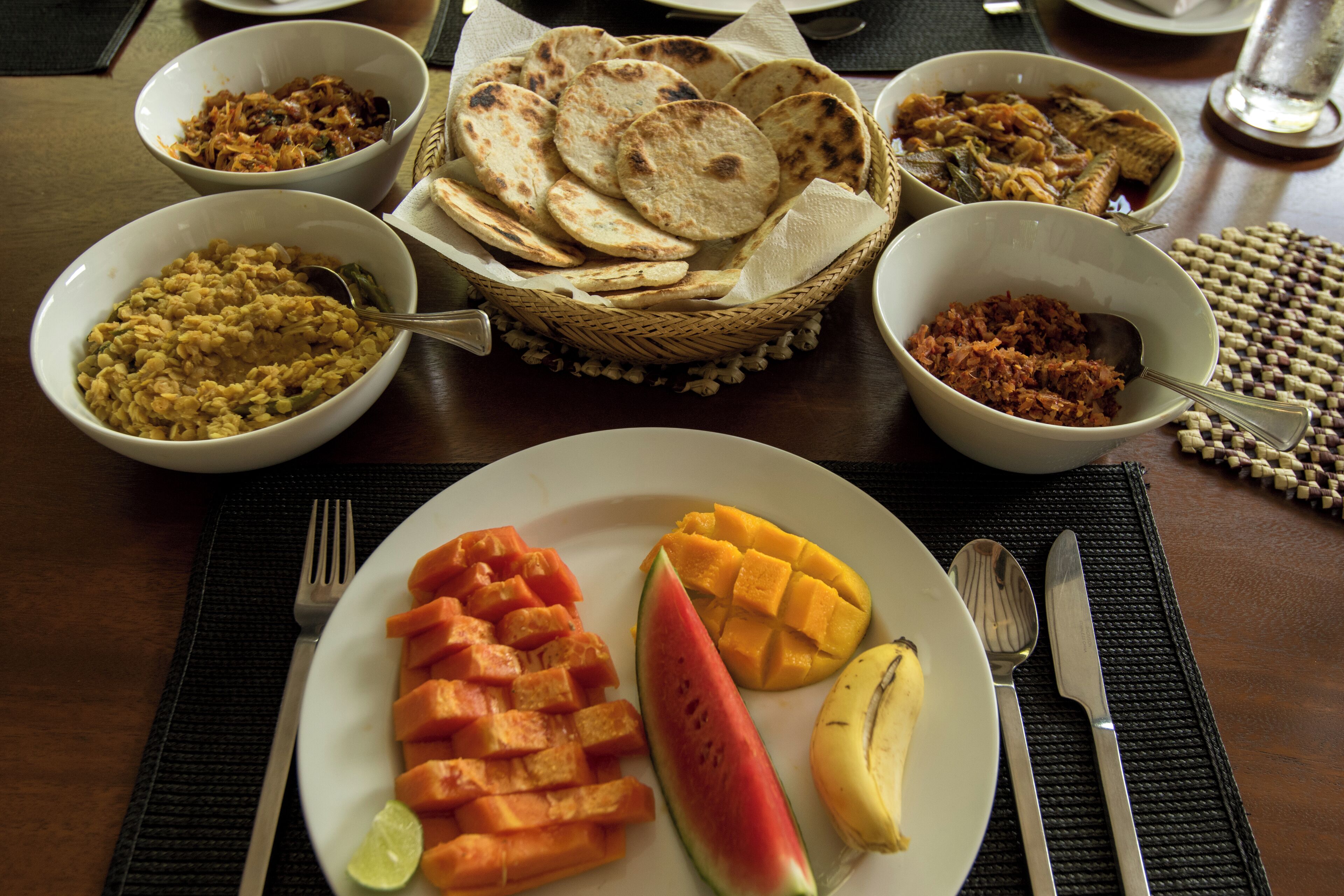 Une table à manger avec divers plats, notamment des galettes, une assiette de mangues, de pastèques et de bâtonnets de carottes, ainsi que des bols contenant différents aliments.