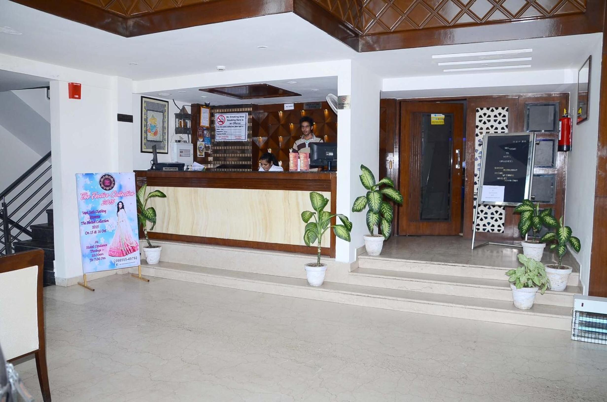 Zona de recepción de un hotel con un mostrador de madera, plantas en macetas, una escalera y un extintor.