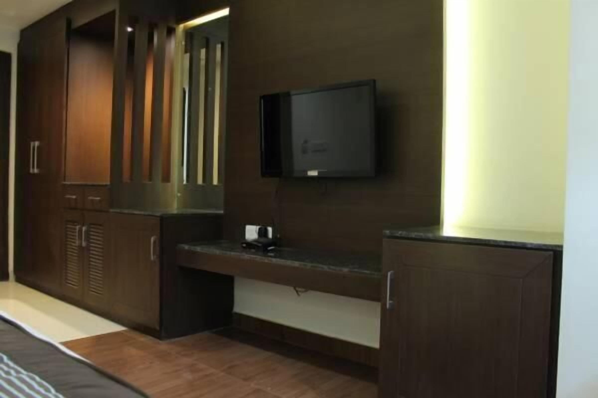 A modern hotel room with a dark wood wardrobe, a flat-screen TV mounted on the wall, and a small desk with a lamp.