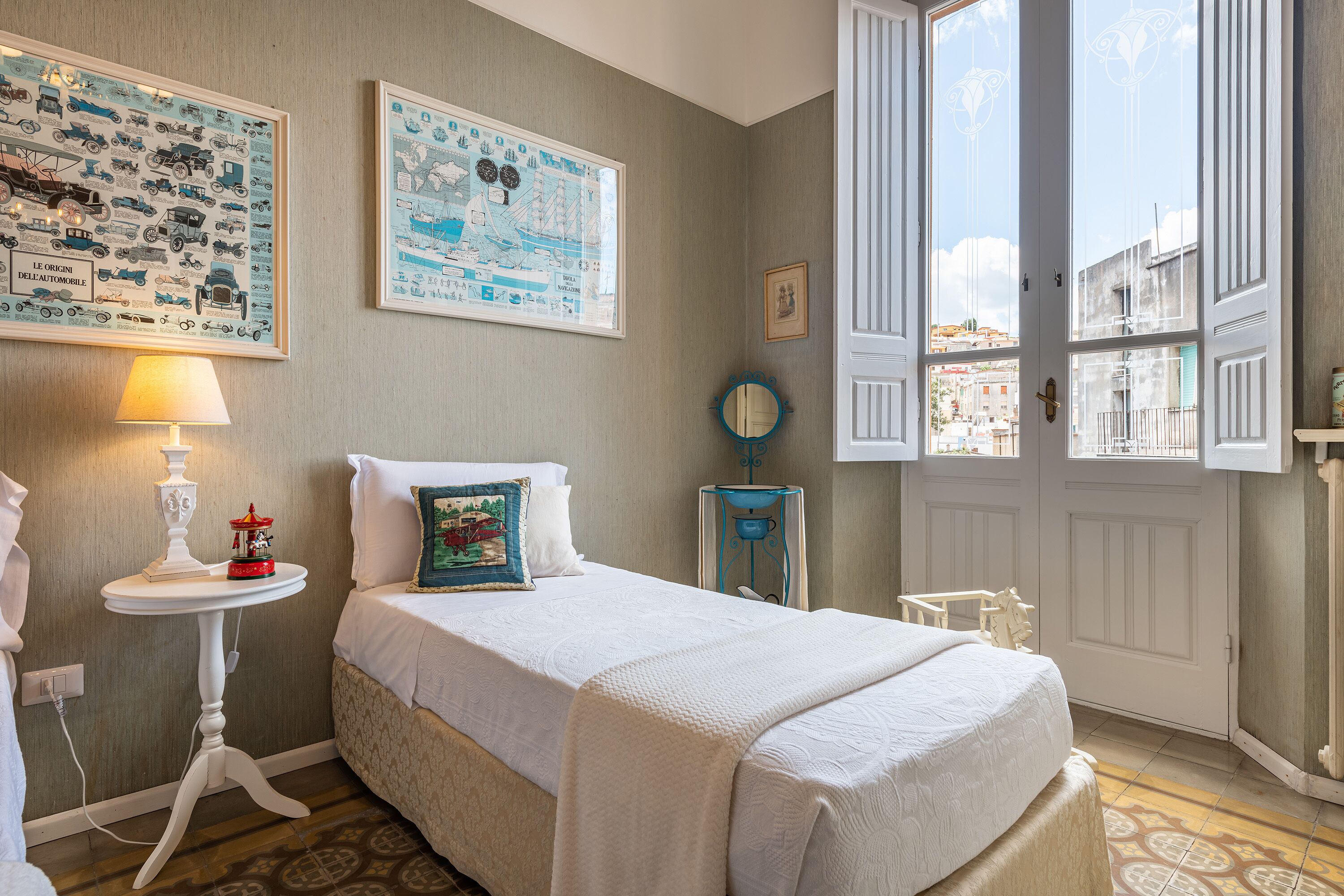 Un dormitorio ordenado con cama, mesita de noche con lámpara, una pequeña mesa con un objeto decorativo y una ventana con vistas a edificios.