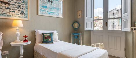 Un dormitorio ordenado con cama, mesita de noche con lámpara, una pequeña mesa con un objeto decorativo y una ventana con vistas a edificios.