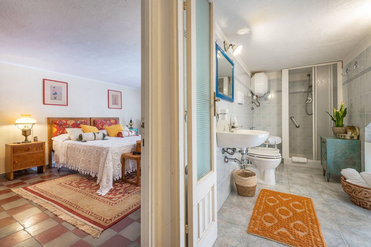 A bedroom with a bed, nightstand, and bedside lamp. Adjacent is a bathroom with a sink, toilet, and shower.
