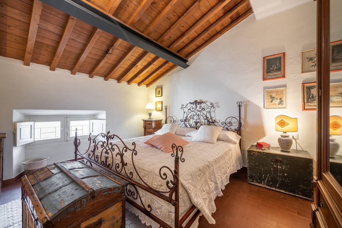 A bedroom with a wooden ceiling, a bed with a metal frame, a dresser, and a window with shutters.