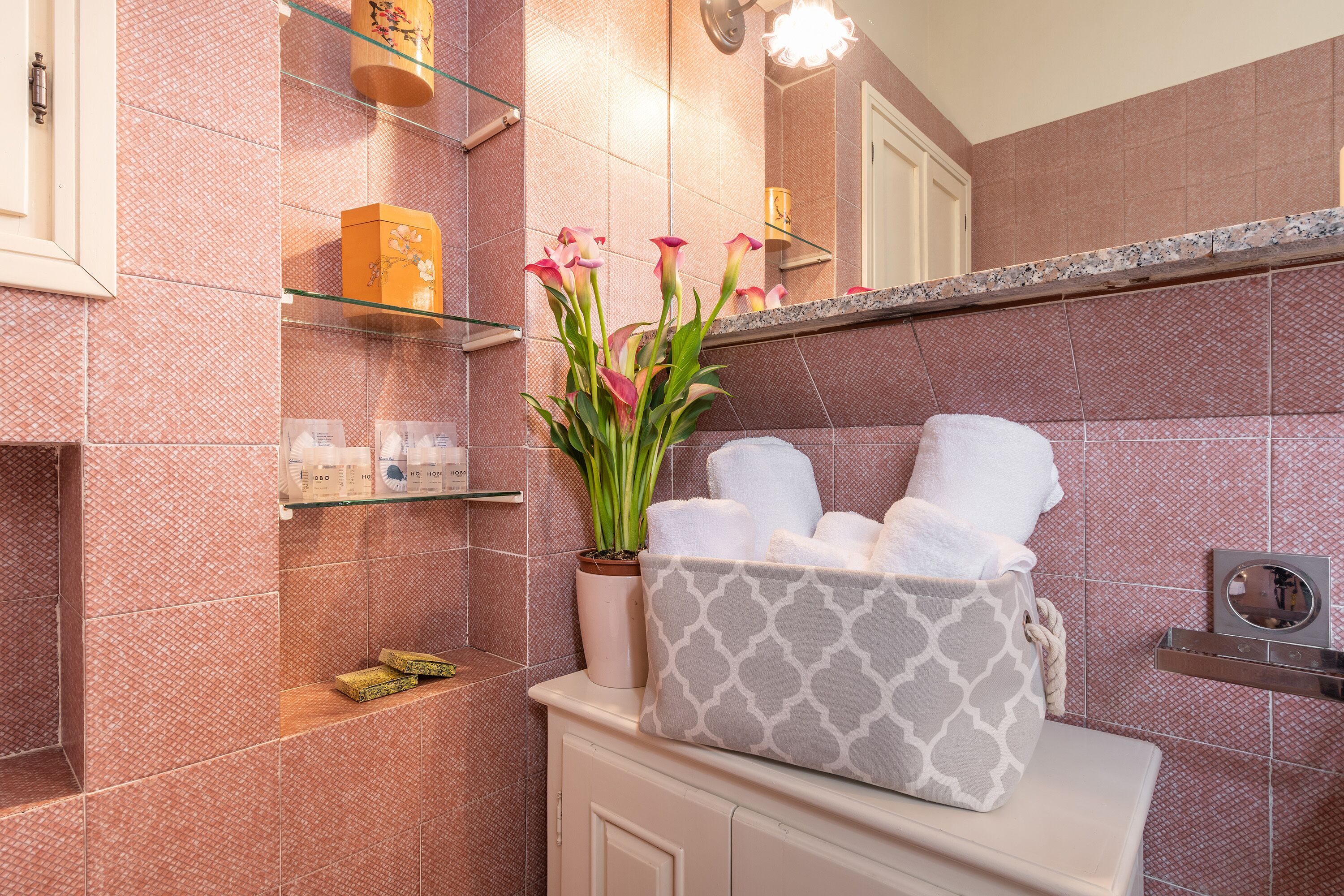 A bathroom with pink patterned tiles, a glass shelf with toiletries, a vase with flowers, and a towel basket.