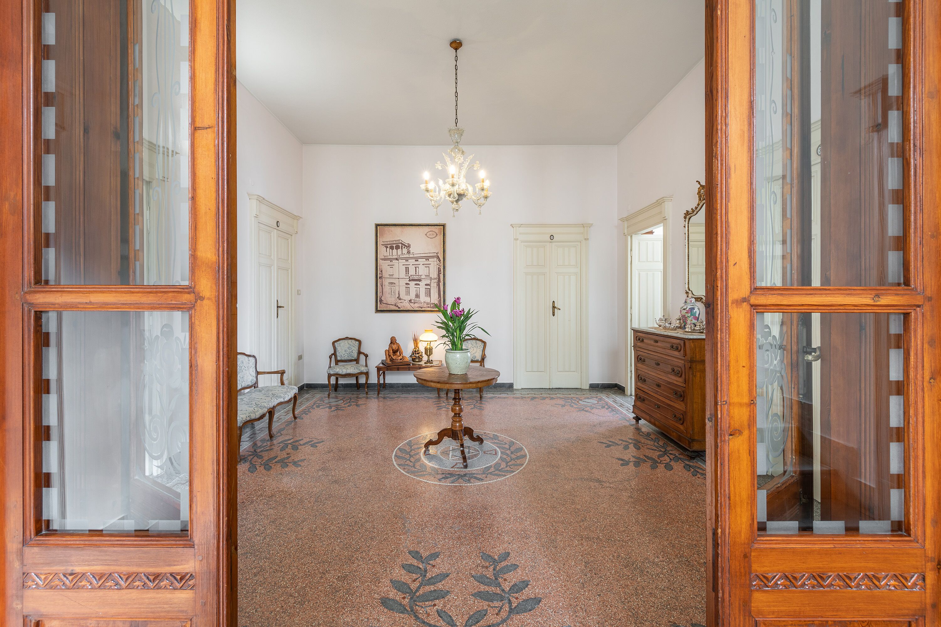 Una habitación amplia con piso estampado, un candelabro, una mesa redonda con un florero y un espejo grande.