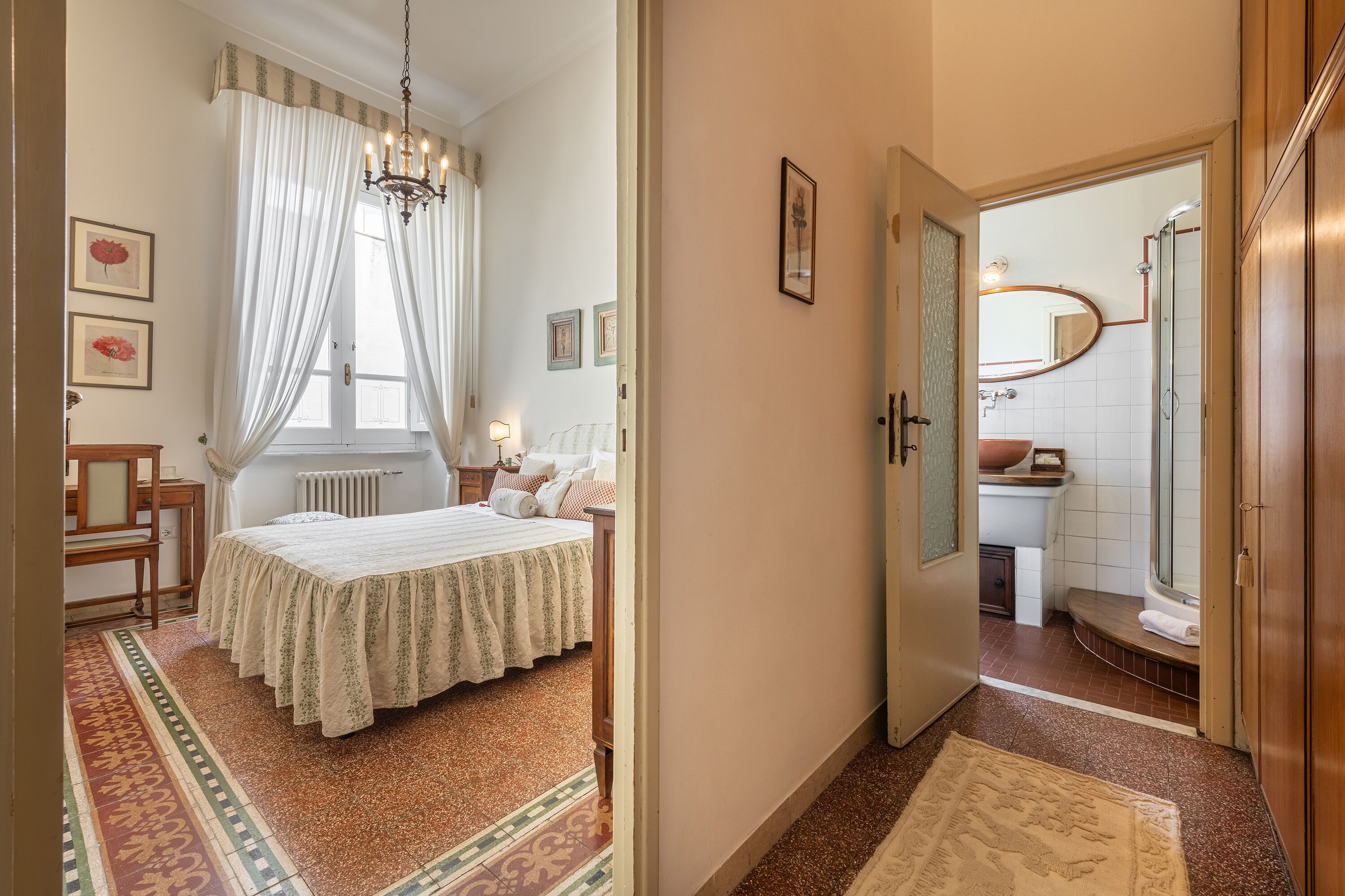 Une chambre à coucher comprenant un lit, une table de chevet, une fenêtre avec des rideaux, un lustre et un couloir menant à une salle de bain équipée d’une douche et d’un lavabo.