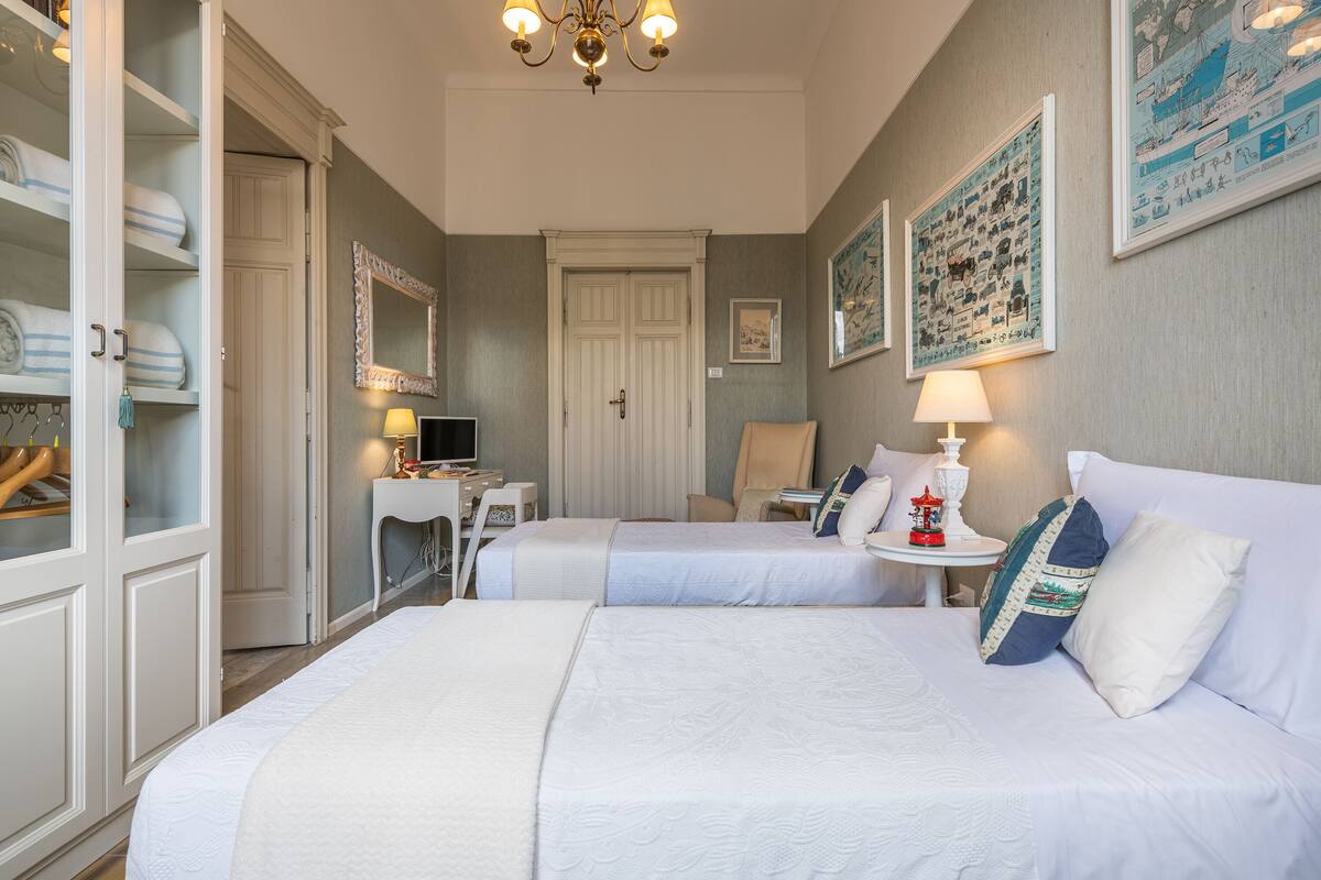A hotel room with two beds, a desk, a chair, a wardrobe, and a chandelier.