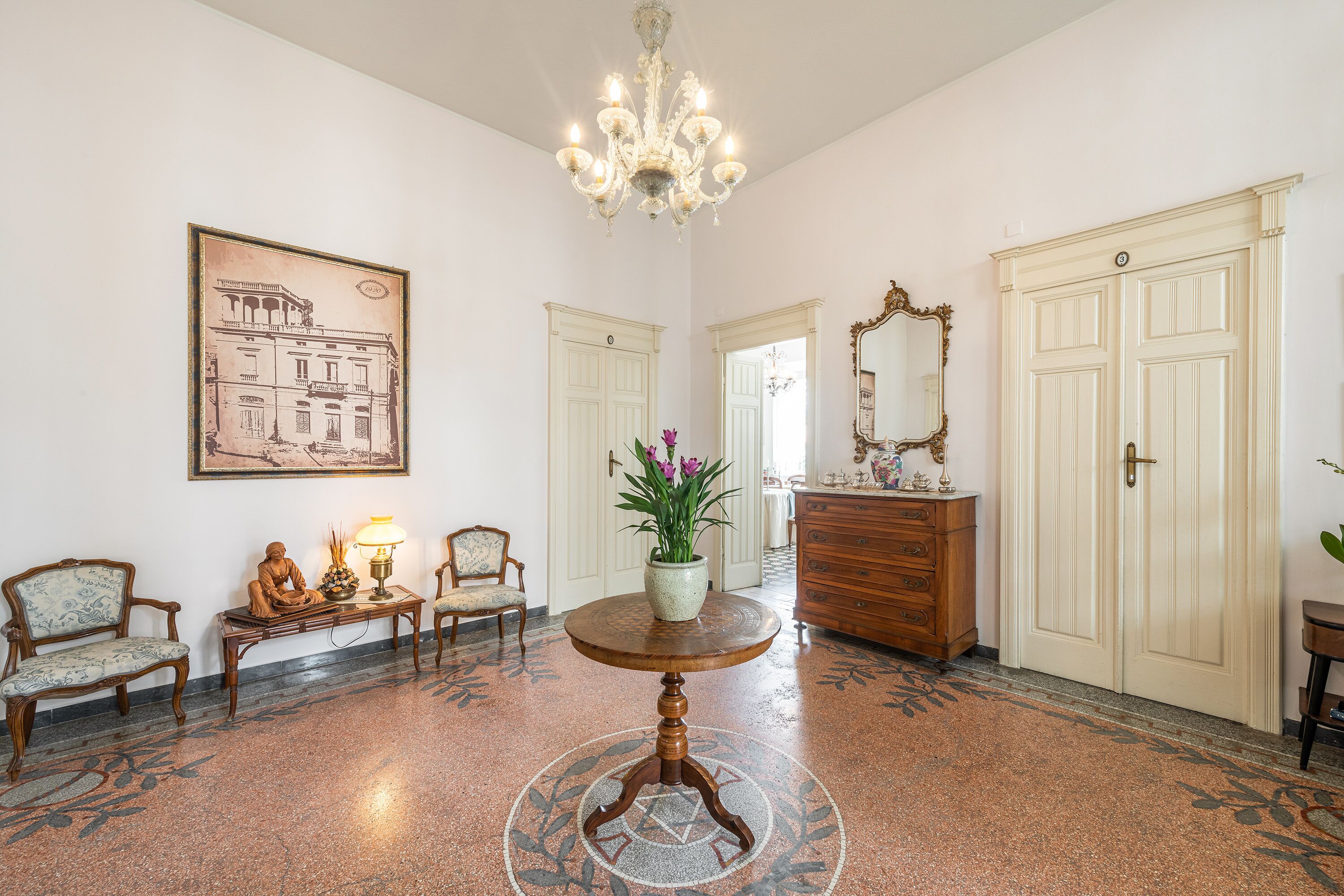A lobby with a round table, chairs, a chandelier, a large mirror, and a door with a number.